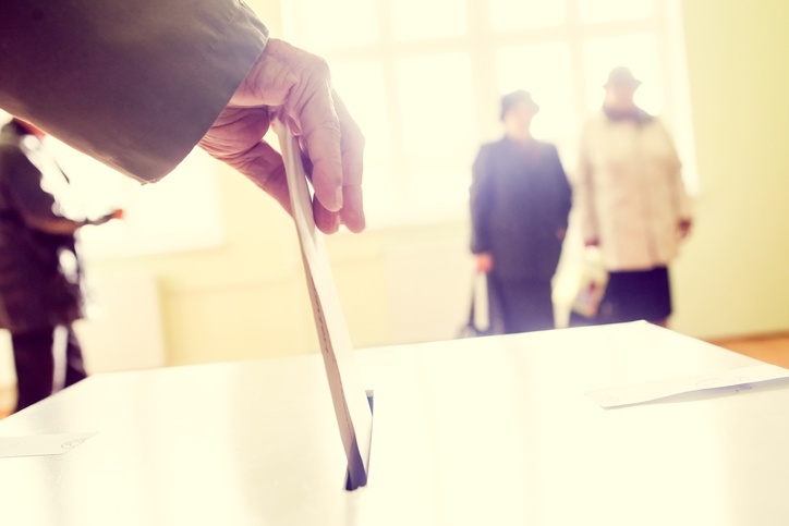 Wähler steckt Wahlzettel in Wahlurne . Im Hintergrund stehen 2 Wähler