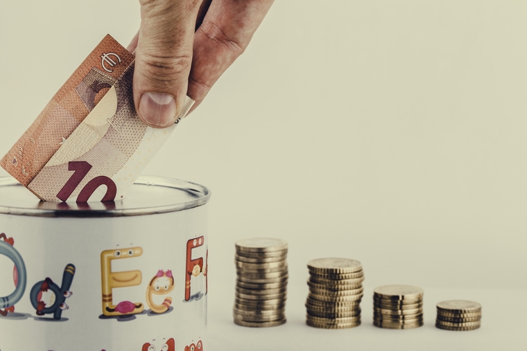 Es ist eine Hand abgebildet, die einen 10 Euro Schein in eine Spardose wirft. Daneben sind Geldmünzen aufgetürmt.